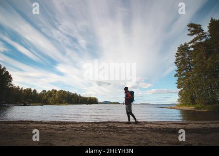 L'escursionista che indossa una giacca e porta uno zaino cammina lungo la spiaggia e guarda il lago Jatkonjarvi al tramonto nel Parco Nazionale di Koli, Finlandia orientale. Ma Foto Stock