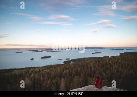 Vista di un avventuriero in camicia rossa e nera seduta sul bordo di una scogliera che guarda il Lago Pielinen nel Parco Nazionale di Koli nella Finlandia orientale. Tramonto ov Foto Stock