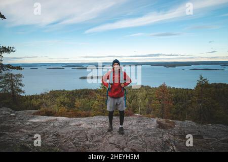 Ridendo zaino in piedi su una roccia con il lago Jatkonjarvi dietro di lui nel Parco Nazionale di Koli, Finlandia orientale. L'uomo, di 24 anni, sorride naturalmente. CA Foto Stock