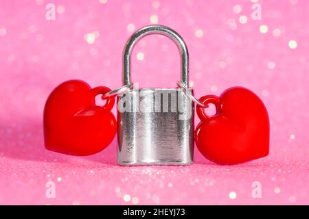 Due ciondoli rossi a forma di cuore attaccati a un lucchetto di tonalità argento su uno sfondo rosa scintillante. Il concetto di amore eterno tra una coppia Foto Stock