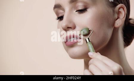 giovane donna con rullo di giada sul volto isolato sul beige Foto Stock