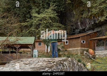 Un uomo vecchio sta preparando legna da ardere nelle montagne della Georgia. Un uomo porta un'armatura di legna da ardere sulla spalla lungo un antico ponte di pietra di fronte Foto Stock