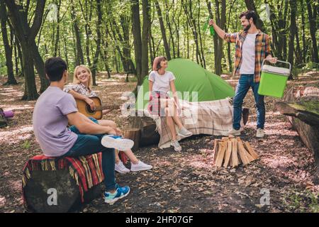 Foto a grandezza naturale dei giovani amici squadra che rimane in campeggio nella foresta estiva suonare la chitarra bere birra Foto Stock