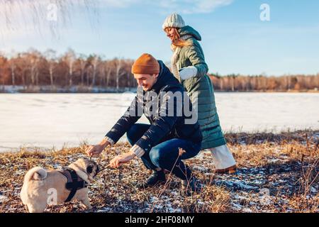 Giovane coppia che cammina con il pug dog nel nevoso parco invernale. Master giocando con il bastone di sostegno dell'animale domestico. Animale mordente ramo di legno all'aperto divertirsi Foto Stock