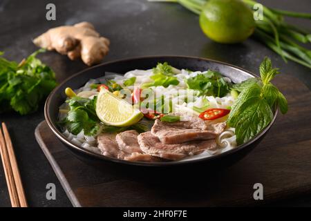 Zuppa tradizionale Pho Bo con manzo, spaghetti di riso, zenzero, lime, peperoncino in ciotola su sfondo nero. Primo piano. Cucina vietnamita e asiatica. Foto Stock