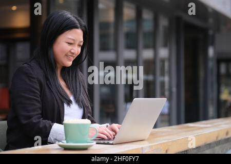Ritratto di sorridente giovane donna asiatica d'affari che lavora su un computer portatile in un bar all'aperto Foto Stock