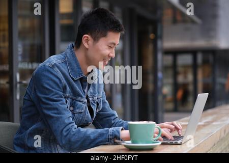 Ritratto di un giovane asiatico seduto al bar all'aperto, sorridente e digitando sul computer portatile Foto Stock