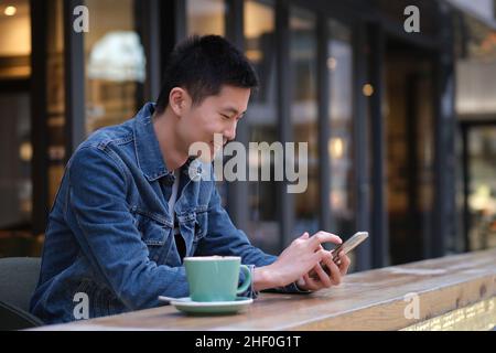 Ritratto di giovane asiatico al bar all'aperto, con telefono cellulare, con sorridere felice Foto Stock