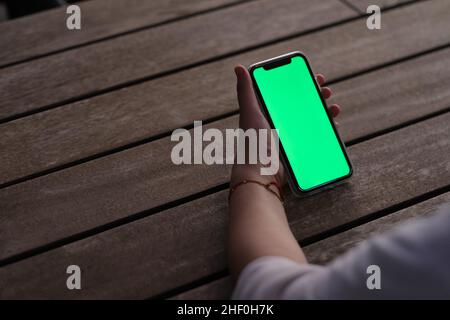 vista sulle spalle del telefono cellulare con schermo verde portatile tenuto a mano su un tavolo da caffe' in legno di notte Foto Stock