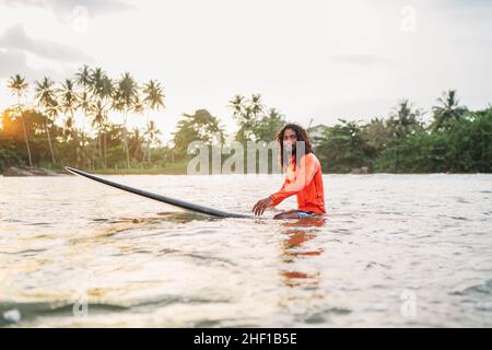 Teen uomo nero dai capelli lunghi che galleggia su una lunga tavola da surf, in attesa di un'onda pronta per il surf con palme boschetto litted al tramonto raggi. Sport acquatici estremi a Foto Stock