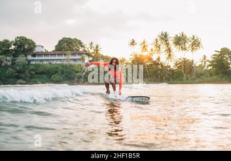 Ragazzo teen nero dai capelli lunghi che guida una lunga tavola da surf. Ha preso un'onda in una baia indiana dell'oceano con sfondo magico del tramonto. Sport acquatici estremi ed es Foto Stock