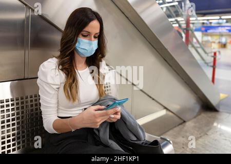 Giovane donna caucasica con maschera medica utilizzando il suo smartphone mentre si aspetta in una stazione della metropolitana. Spazio per il testo. Foto Stock