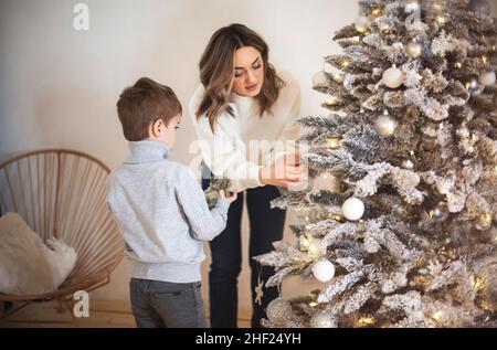 Bambino piccolo di preschool che aiuta la madre a decorare l'albero di Natale nel paese, la mamma ed il figlio piccolo che decorano insieme il firtree di natale, il genitore giovane della famiglia ed il ch Foto Stock