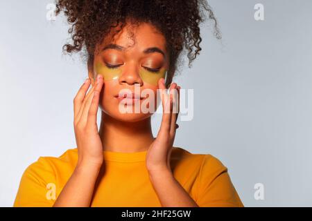 Concetto di cura della pelle. Giovane calma afroamericana donna godendo di routine di bellezza, applicazione di idrogel cosmetici occhi patch per la zona sotto-occhio sul viso, isolare Foto Stock