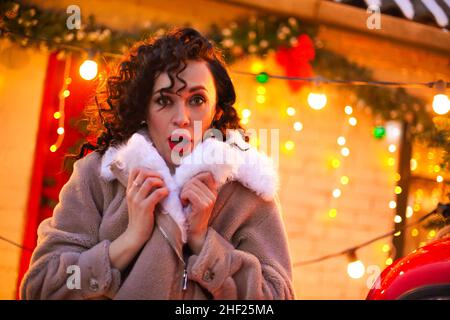 Donna di Natale sorpresa con trucco festivo e acconciatura guardando la macchina fotografica con volto stupito, in piedi all'aperto contro casa di mattoni decorata con x Foto Stock