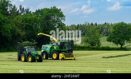 Trattori haymaking, impegnati nella guida in campo agricolo con la trebbiatrice John Deere, carico di rimorchi di riempimento (insilato da erba tagliata) - Yorkshire Inghilterra UK. Foto Stock