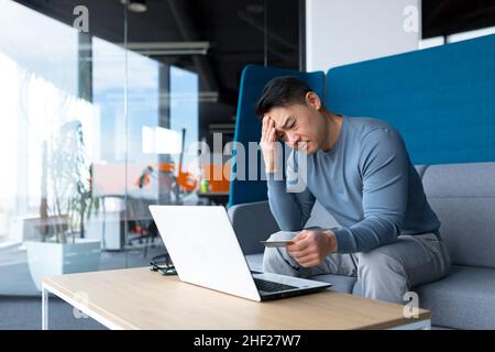 L'uomo asiatico infastidito che prova a fare le operazioni bancarie con il laptop e la carta di credito, uomo d'affari nel lavoro nell'ufficio moderno Foto Stock