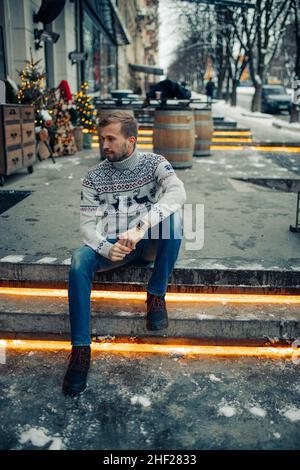 Il giovane uomo si siede sulle scale e guarda con cura alla strada decorata per Natale. Foto Stock