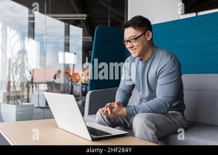 Asian con gli occhiali, lavorare in ufficio, fare una videochiamata con un laptop, sorridendo e gioendo, comunicare in remoto con i colleghi online, bu Foto Stock