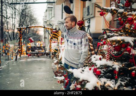 Il giovane uomo si alza e sorride vicino all'edificio in una strada innevata decorata per Natale. Foto Stock