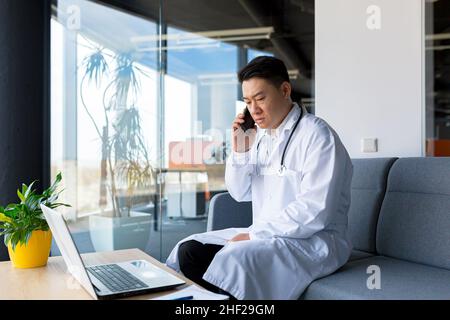 Il medico asiatico sconvolto segnala cattive notizie al paziente, seduto in clinica, uomo che parla al telefono Foto Stock