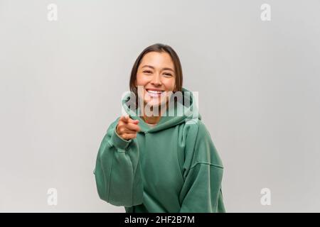 Donna felice soddisfatto con capelli marroni puntando le dita su di voi guardando la macchina fotografica con un sorriso toothy, congratulandosi con la vittoria. Studio interno girato isolato su sfondo bianco Foto Stock