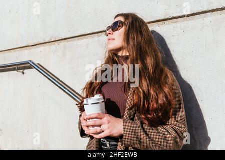 Angolo di donna basso con tazza di caffè ecologico da asporto appoggiata su pareti in cemento e guardando via nella giornata di sole in città Foto Stock