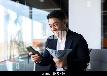 Felice riuscito imprenditore asiatico conta, onde, tiri, soldi in ufficio guardando la macchina fotografica Winner successo nel mercato azionario delle scommesse che celebra la vittoria Foto Stock