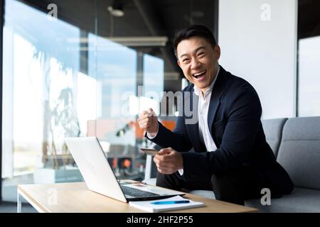 Felice uomo d'affari asiatico che trasferisce la carta di credito, felice e sorridente uomo che acquista in negozio online seduto in ufficio Foto Stock