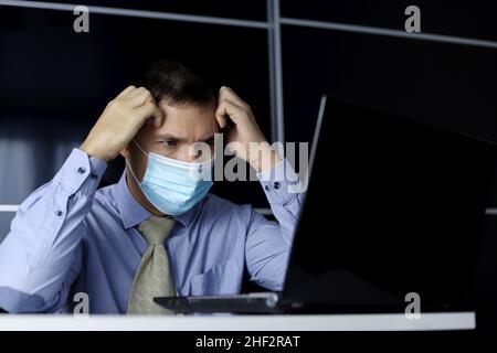 Uomo in maschera facciale e abiti da ufficio che guarda sconvolto al display del portatile. Concetto di risoluzione di un'attività difficile, errore critico Foto Stock