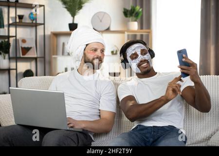 Cura della pelle degli uomini a casa. Due giovani uomini multietnici che fanno le procedure di igiene del viso, utilizzando le patch per gli occhi e la maschera facciale fare selfie foto sullo smartphone. Coppia gay a casa Foto Stock