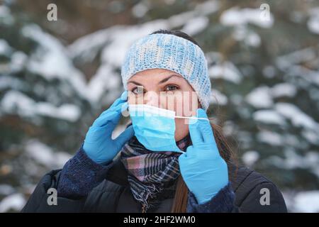 Giovane donna in caldo inverno vestiti, indossare guanti di protezione, mettere o rimuovere monouso monouso blu viso virus bocca maschera, closeup, sfocato Foto Stock