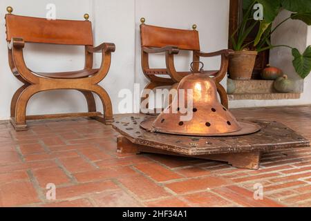 cortile interno scena con un antico tavolo braciere su un piedistallo in legno di fronte a due sedie in legno antico-stile. il braciere è stato utilizzato per molti Foto Stock