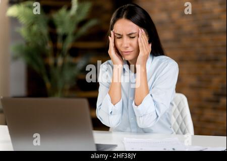 Sopraffarato triste caucasica donna d'affari, manager o segretario, stanco di lavorare a un computer portatile, stanco di sedersi al suo posto di lavoro in ufficio, massaggiando le sue tempie, chiudendo gli occhi, ha bisogno di riposo Foto Stock
