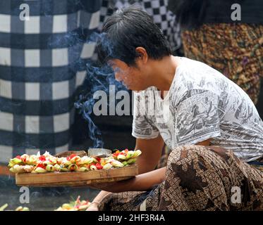 Un giovane uomo balinese si accoccolò e trasportò un vassoio di rattan con offerte hindu canang sari nel mercato di Sukawati a Bali, Indonesia Foto Stock