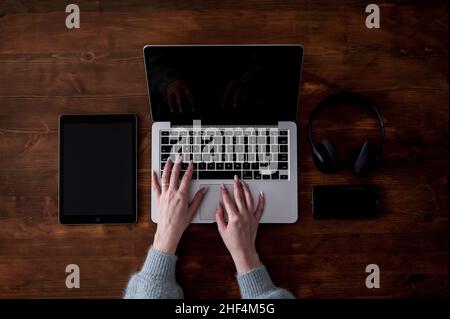 Un computer portatile, un tablet, un telefono e un paio di cuffie disposte su un tavolo di legno marrone scuro Foto Stock