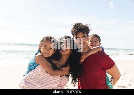 Felice padre biraciale e madre che danno giri di piggyback ai bambini in spiaggia contro il cielo con spazio copia Foto Stock