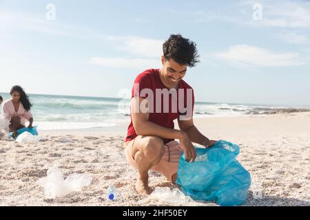 Sorridente uomo biraciale raccolta rifiuti in sacchetto di plastica blu dalla sabbia alla spiaggia contro il cielo Foto Stock