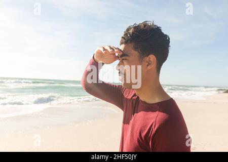 Vista laterale dell'uomo biraciale che protegge gli occhi mentre si guarda via la spiaggia in giorno di sole Foto Stock