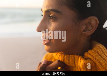 Primo piano di una giovane donna biraciale pensierosa in un maglione che guarda via la spiaggia durante il tramonto Foto Stock