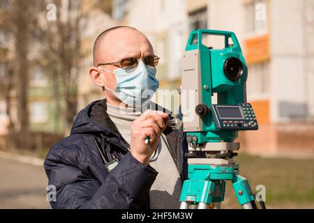 Surveyor in una maschera protettiva funziona con una stazione totale. Concetto di protezione da coronavirus Foto Stock