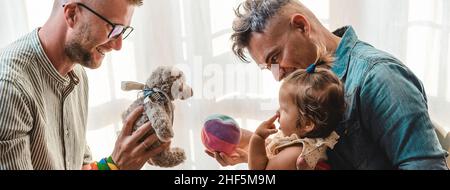 Banner o header orizzontale con coppia gay maschile con baby girl adottata a casa - due padri che giocano con la loro figlia - famiglia LGBTQ+ a casa - di Foto Stock