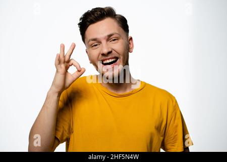 Il ragazzo allegro in una T-shirt gialla mostra un gesto di Okey su uno sfondo bianco nello Studio. Gesto positivo, gesto OK. Spazio copia. Primo piano. Foto di alta qualità Foto Stock