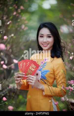 Ho Chi Minh City, Vietnam: Ragazza vietnamita che indossa ao dai in posa con fiori di pesca per celebrare il nuovo anno Foto Stock