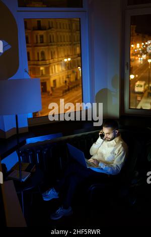 Un dipendente maschio effettua una telefonata mentre lavora durante la notte in ufficio. Programmatore o designer occupato con un computer portatile Foto Stock