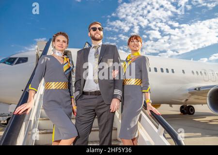Capitano dell'aviazione maschile e due assistenti di volo donne in piedi sulle scale di imbarco aereo e sorridenti Foto Stock