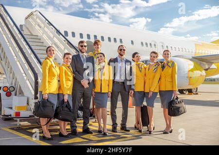 Piloti maschi e assistenti di volo femminile in uniformi aeronautiche guardando la macchina fotografica e sorridendo mentre si trova vicino aereo in campo aereo Foto Stock