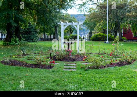 Un arco in legno bianco d'epoca e un recinto di picket in legno bianco circondano un giardino. Il giardino ha lussureggianti alberi verdi e erba verde nel parco. Foto Stock