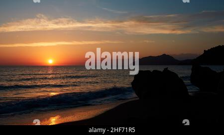 Grecia, Isole greche, Isole IONIE, Corfù, costa occidentale, Agios Gordios, serata scatto, tramonto, blocco dalla costa ad ovest, a sinistra il sole scende, il sole sul mare leggermente commovente, sulle rocce di destra, cielo da rosso arancio a blu, nuvole grigio Bianco Foto Stock