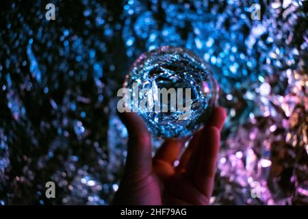 La mano delle donne che tiene una sfera di cristallo del cassiere di fortuna e che guarda nel futuro. Sfondo sfocato e luminoso, sfondo mostrato attraverso il riflesso Foto Stock